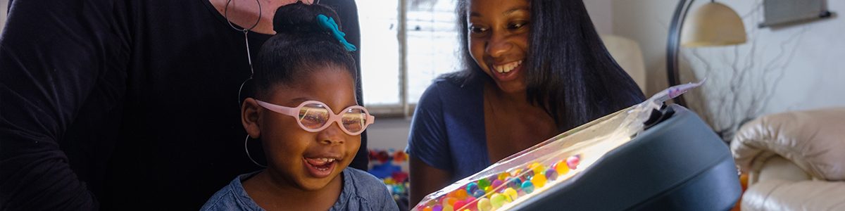 A toddler with glasses smiles as she gazes at a lighbox. Her mother looks on, also smiling.