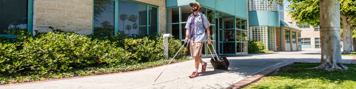 An adult who is blind walks down the sidewalk of Wayfinder's Los Angeles campus, holding a white cane in one hand and a suitcase in the other