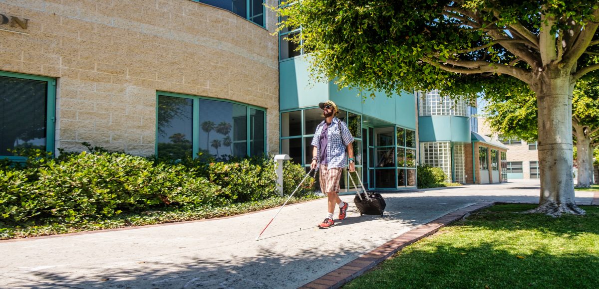 An adult who is blind walks down the sidewalk of Wayfinder's Los Angeles campus, holding a white cane in one hand and a suitcase in the other