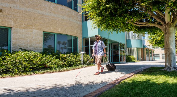 An adult who is blind walks down the sidewalk of Wayfinder's Los Angeles campus, holding a white cane in one hand and a suitcase in the other