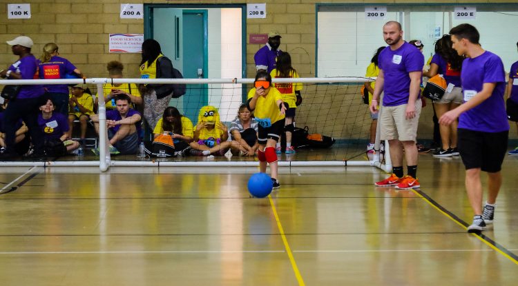 A girl wearing a blindfold throws a blue goalball across a court