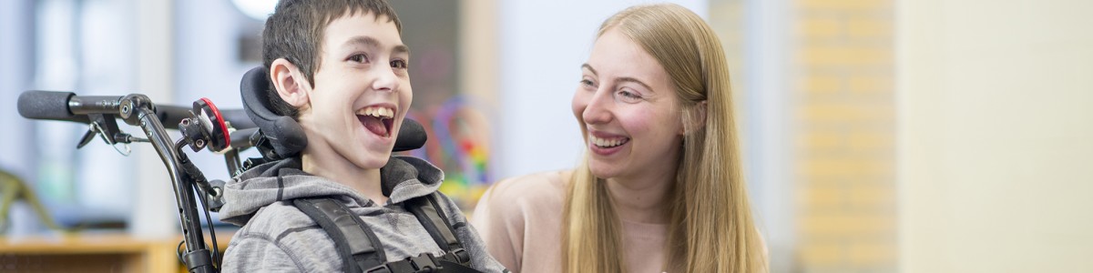 A caregiver is playing with a little boy that has disabilities. He is happily sitting in his wheelchair.