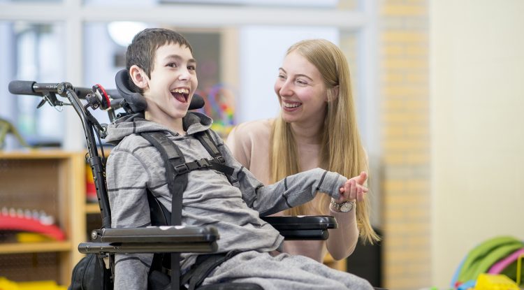 A caregiver is playing with a little boy that has disabilities. He is happily sitting in his wheelchair.