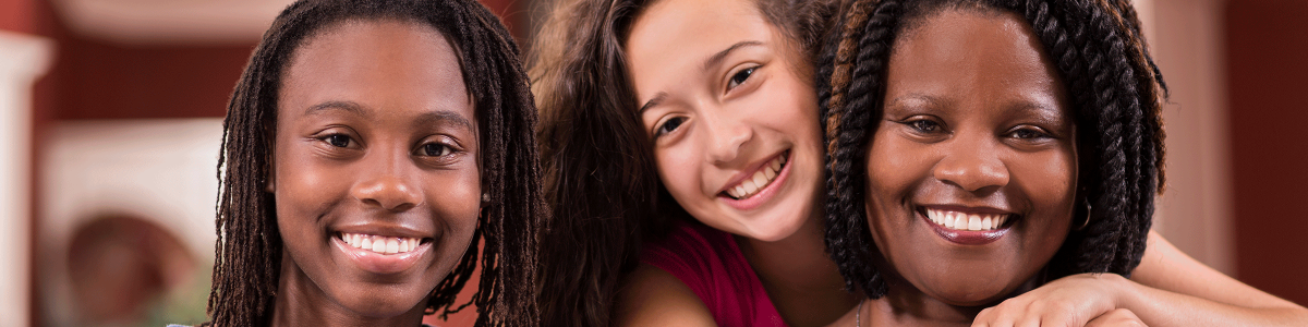 A family of a mother and two young daughters smiles at the camera. One of the daughters is standing behind her mother with her arms around her shoulders.