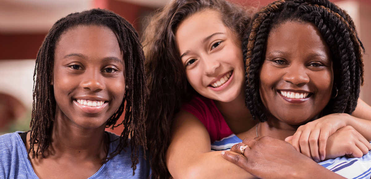 A family of a mother and two young daughters smiles at the camera. One of the daughters is standing behind her mother with her arms around her shoulders.