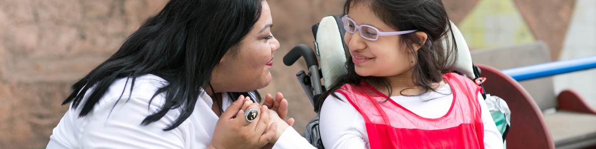 A staff of Wayfinder's special education / special needs school kneels next to a student in a wheelchair and holds her hand