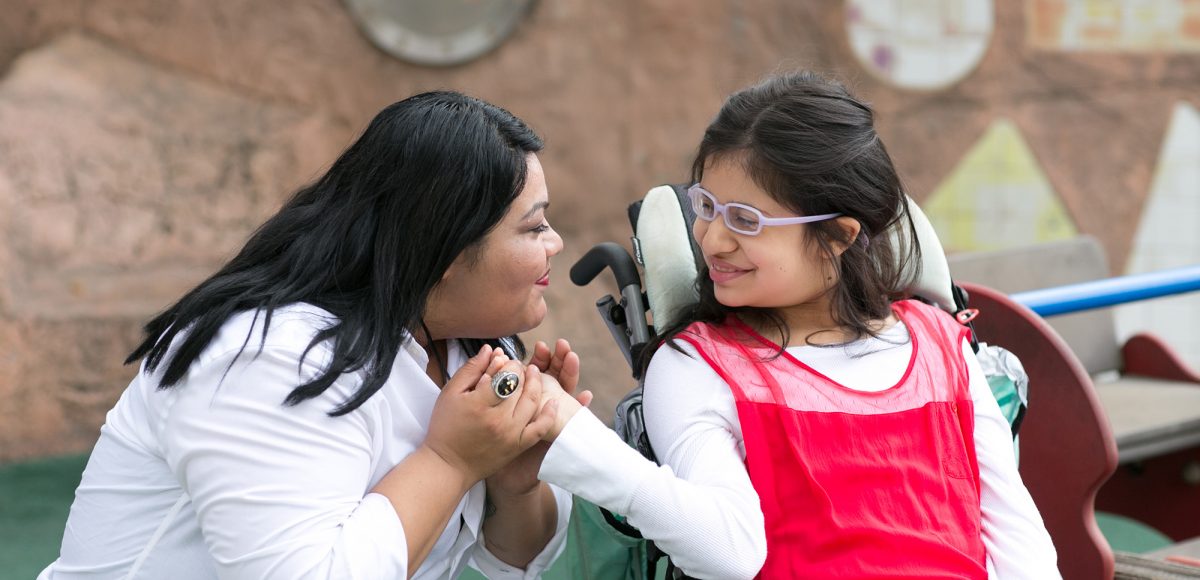A staff of Wayfinder's special education / special needs school kneels next to a student in a wheelchair and holds her hand
