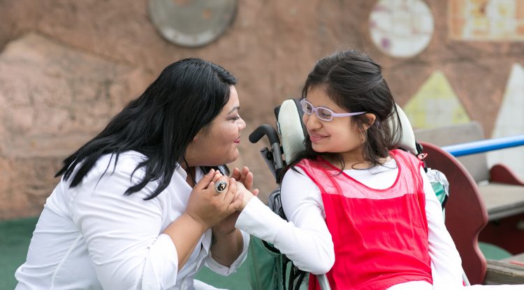 A staff of Wayfinder's special education / special needs school kneels next to a student in a wheelchair and holds her hand