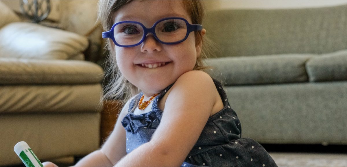 A toddler wearing glasses smiles at the camera.