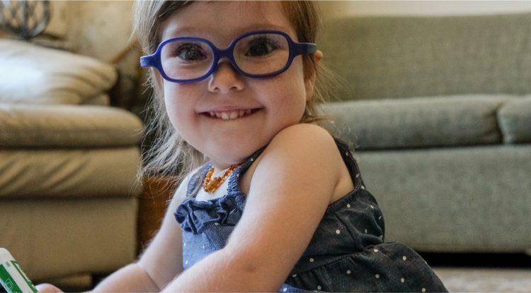 A toddler wearing glasses smiles at the camera.