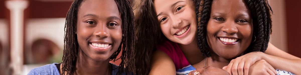 A family of a mother and two young daughters smiles at the camera. One of the daughters is standing behind her mother with her arms around her shoulders.