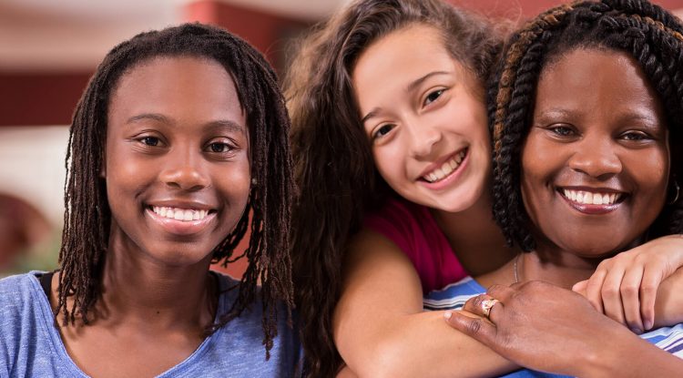 A family of a mother and two young daughters smiles at the camera. One of the daughters is standing behind her mother with her arms around her shoulders.