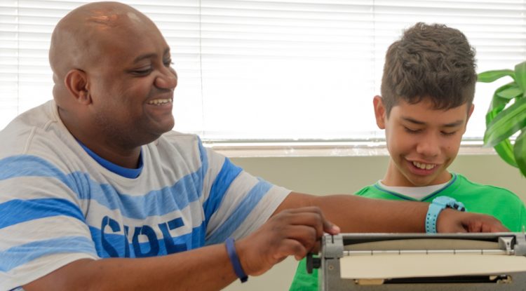 An aide assists a student with his Braille machine as the student smiles