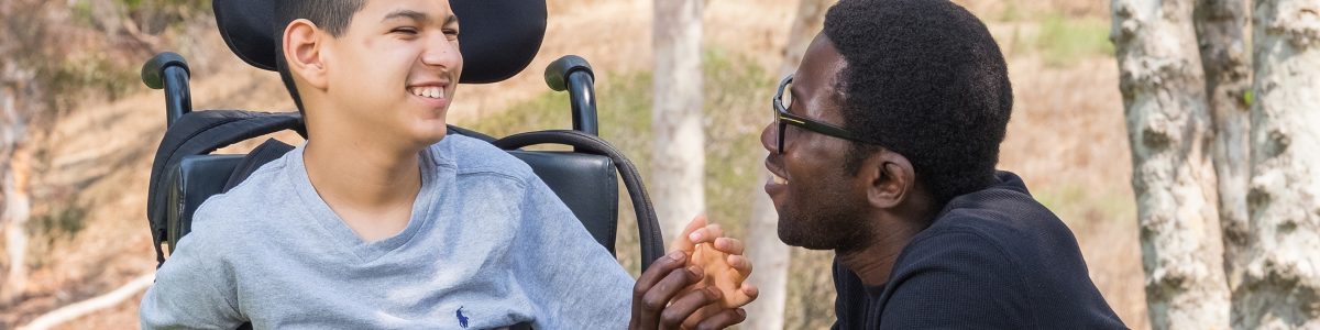 An aide kneels next to a young boy in a wheelchair. They are holding hands and smiling at one another.