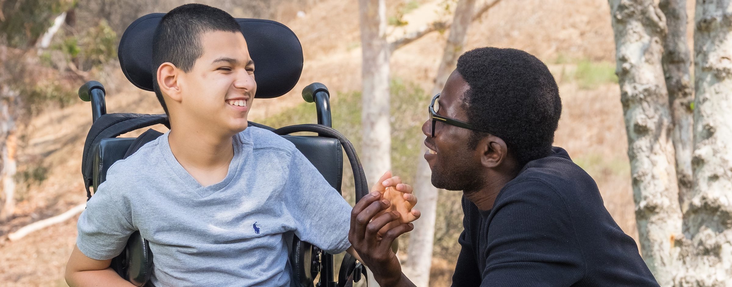 An aide kneels next to a young boy in a wheelchair. They are holding hands and smiling at one another.