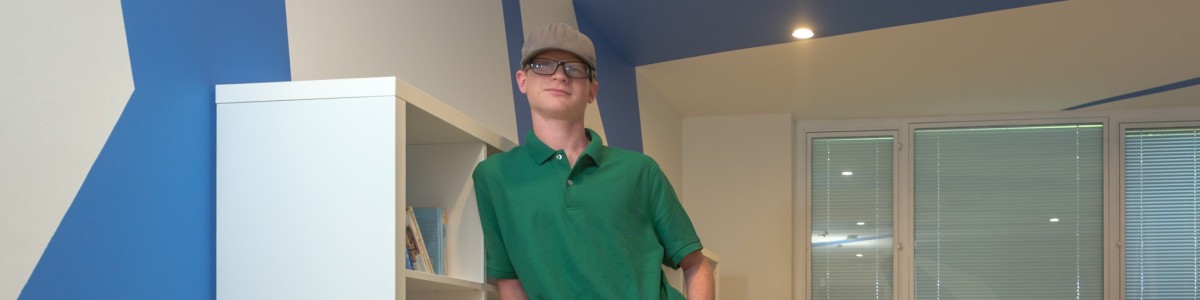 A young man with glasses and a hat leans against a bookshelf, smiling at the camera