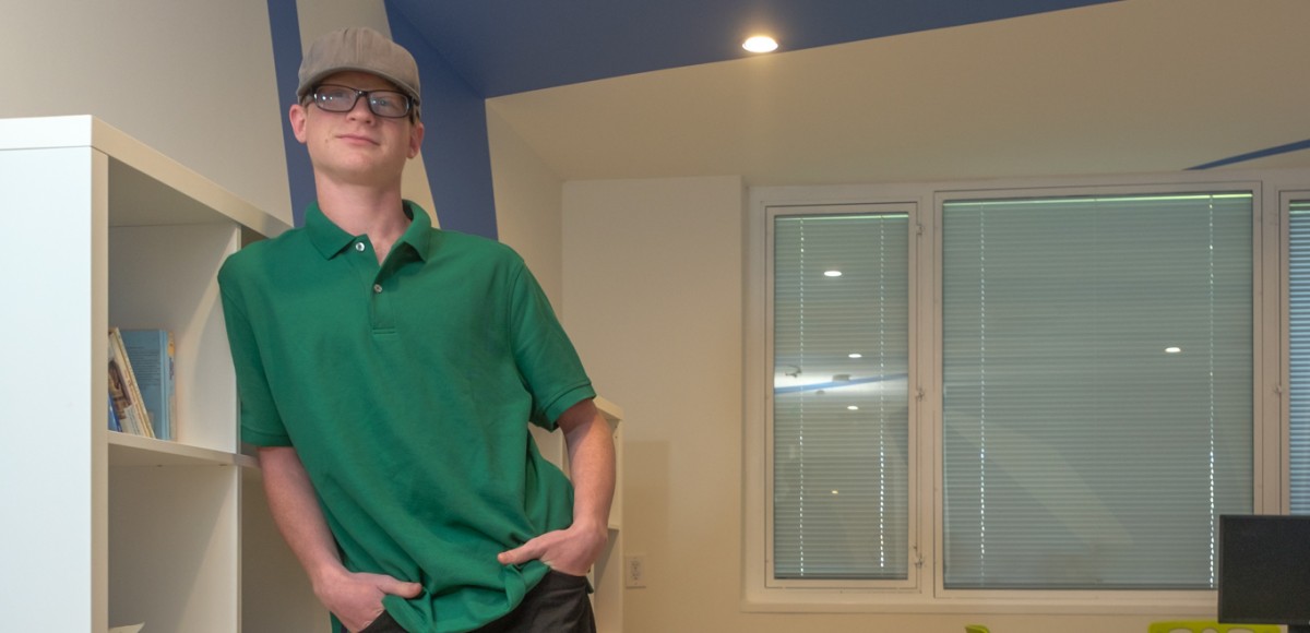 Un joven con gafas y sombrero se apoya en una estantería, sonriendo a la cámara.