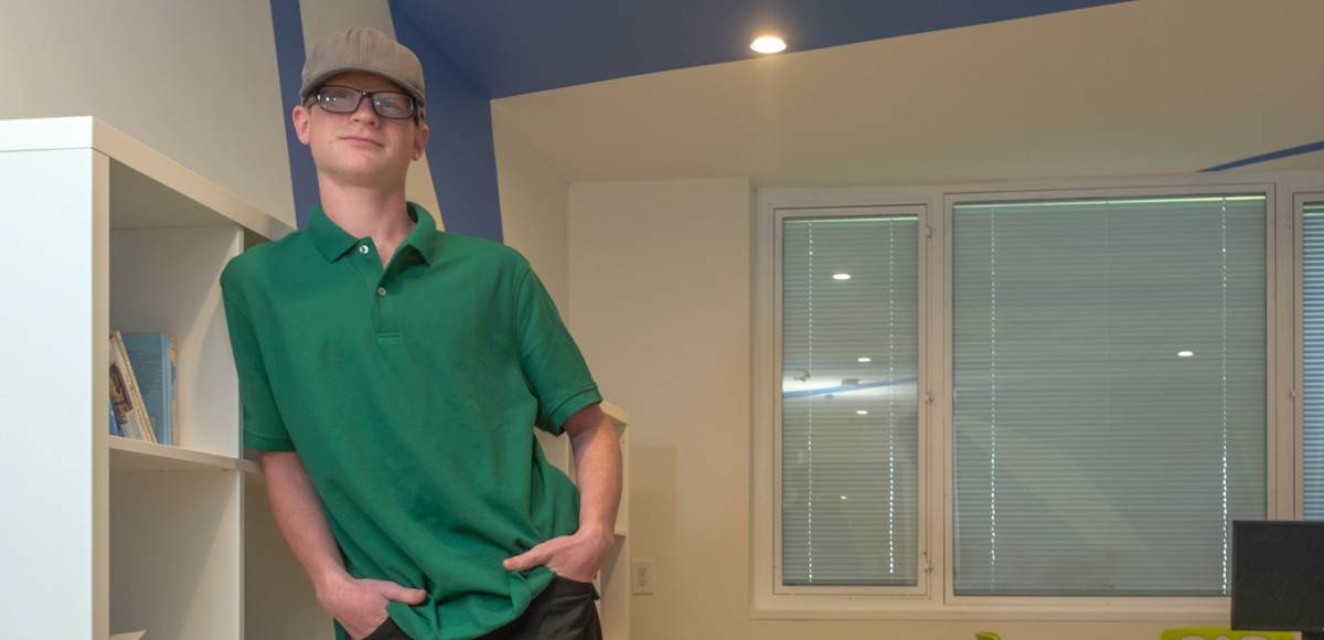 A young man with glasses and a hat leans against a bookshelf, smiling at the camera