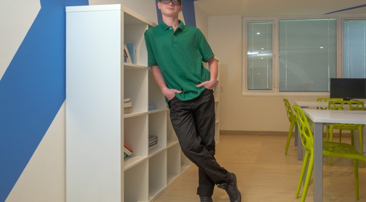 A young man with glasses and a hat leans against a bookshelf, smiling at the camera