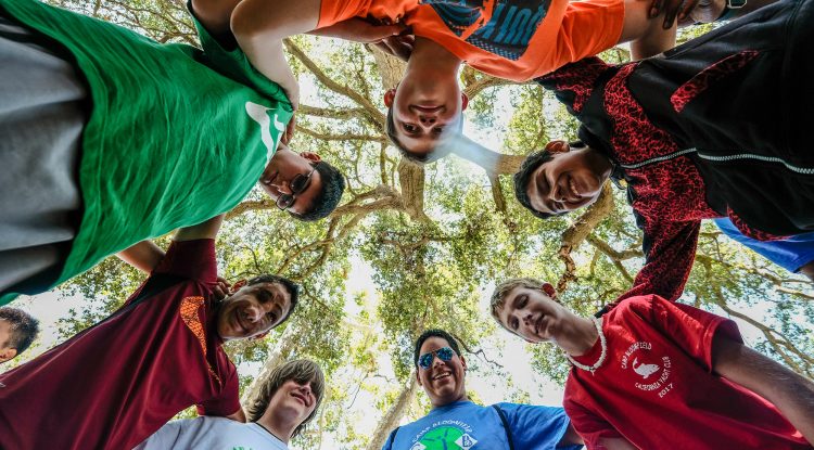 A group of boys with their arms around one anothers' shoulders stand in a circle, smiling down at the camera at Camp Bloomfield.