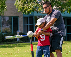 An instructional aide assists a young boy who is visually impaired to hit a ball on a tee with a bat