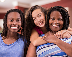 A family of a mother and two young daughters smiles at the camera. One of the daughters is standing behind her mother with her arms around her shoulders.