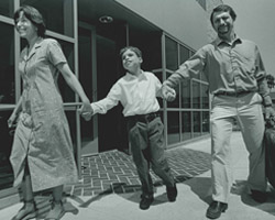 A child walks between his mother and father, smiling, on the campus of Junior Blind. The father is holding a suitcase.