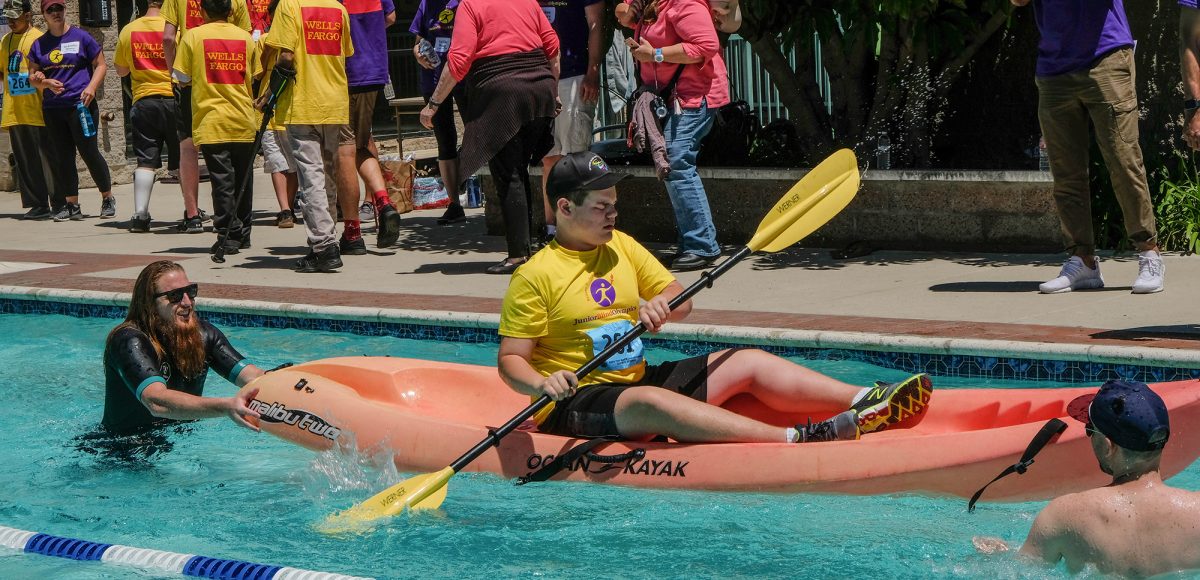 A young boy who is visually impaired paddles a kayak, guided by two volunteers at the Wayfinder Paralympic Games