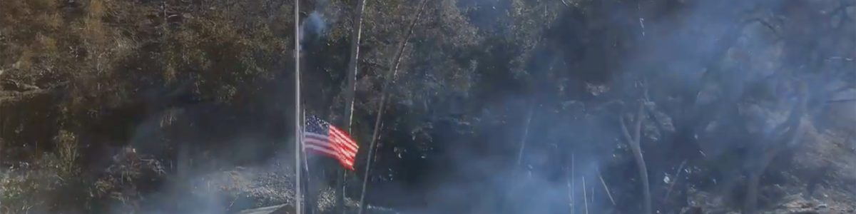 The American flag flying at half-staff with smoky remains of Camp Bloomfield