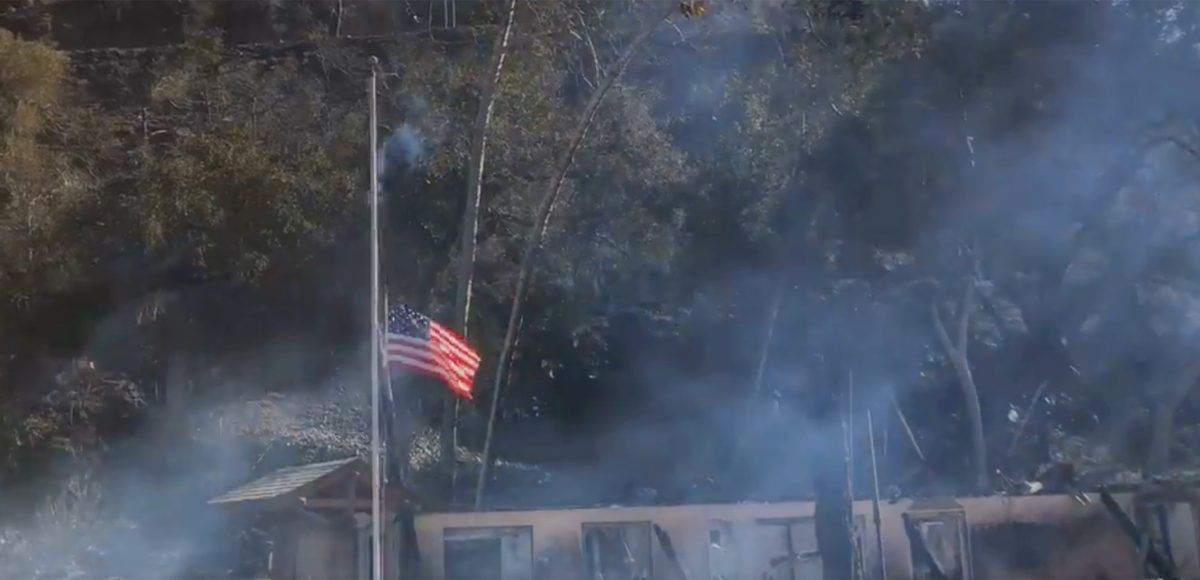 The American flag flying at half-staff with smoky remains of Camp Bloomfield