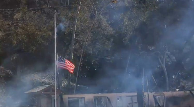 The American flag flying at half-staff with smoky remains of Camp Bloomfield