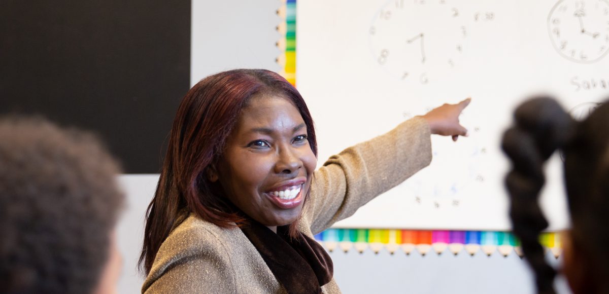 smiling woman points to white board in classroom while students watch