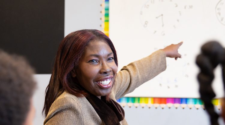 smiling woman points to white board in classroom while students watch