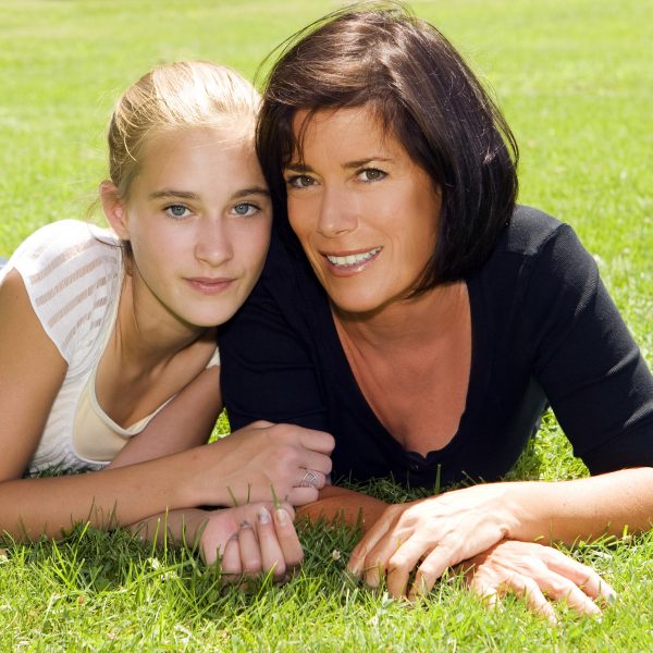 blond teenage girl and brunette mother smile while posing on grass