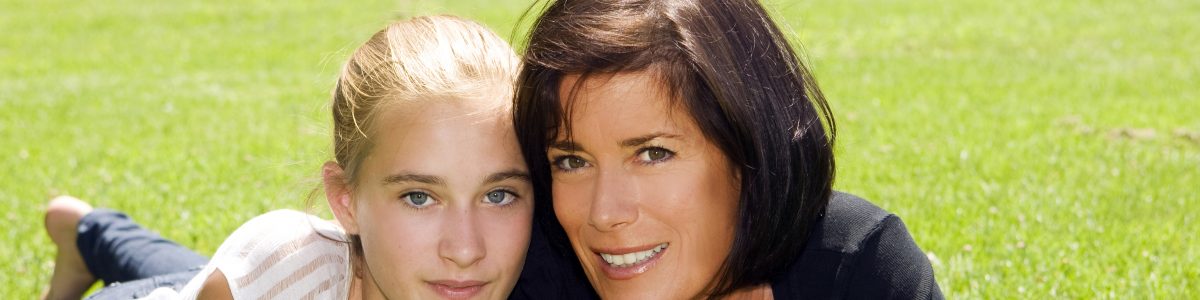 blond teenage girl and brunette mother smile on grassy area