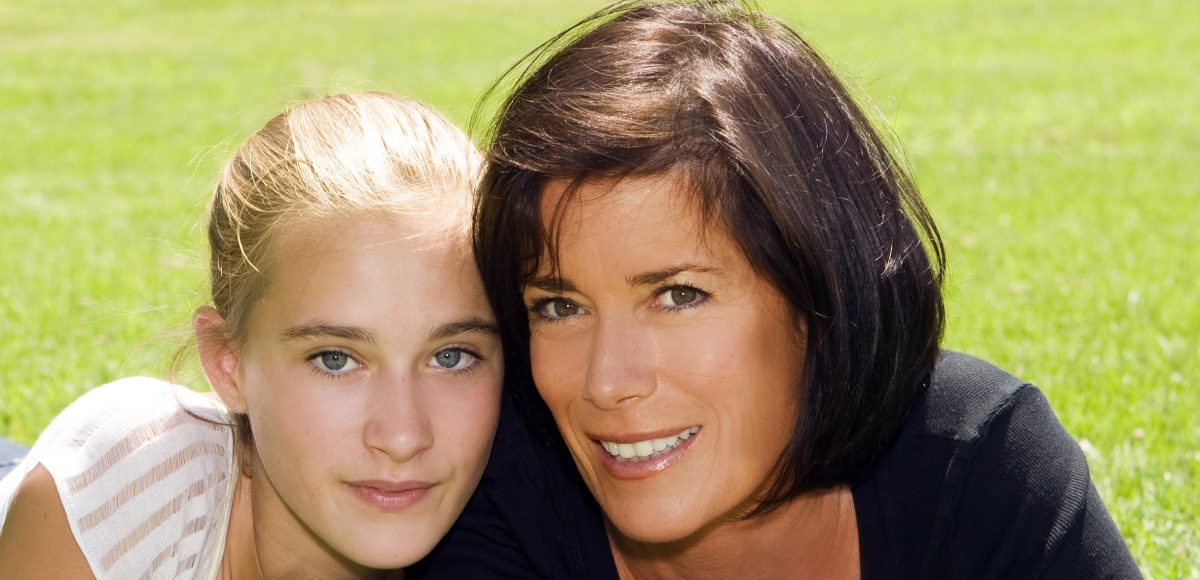 blond teenage girl and brunette mother smile on grassy area