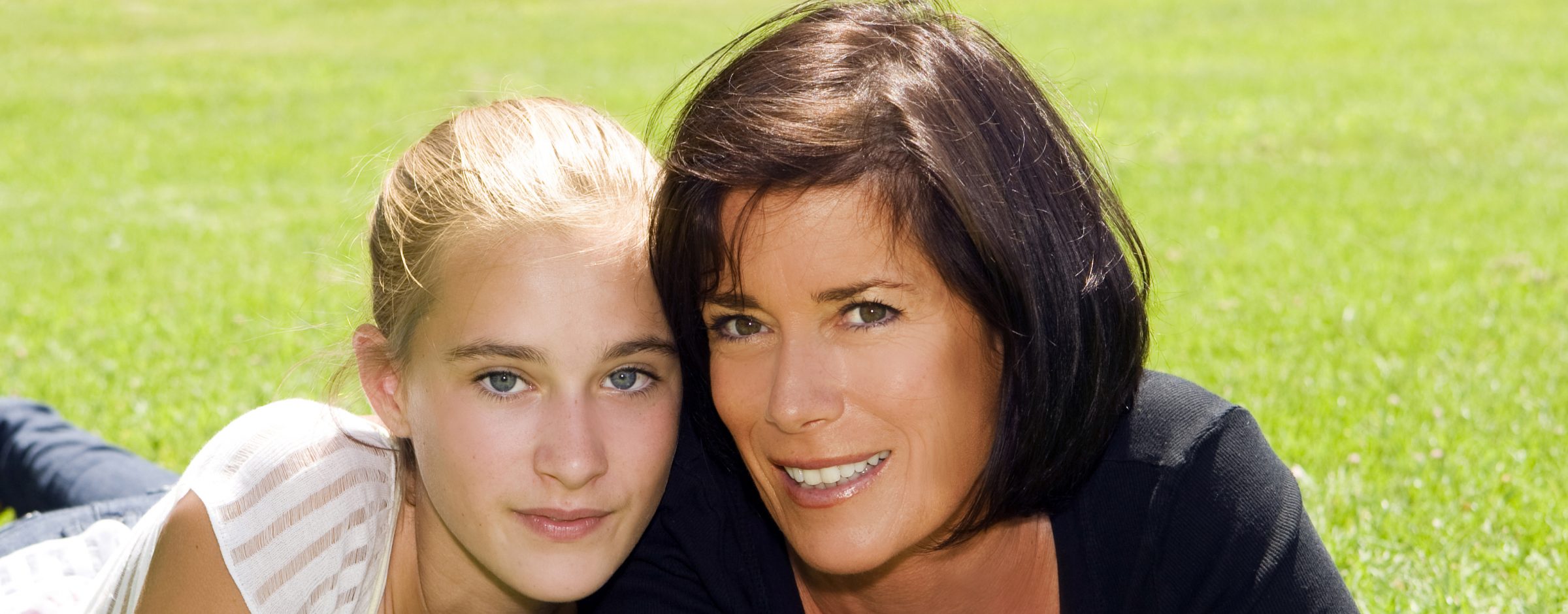 blond teenage girl and brunette mother smile on grassy area
