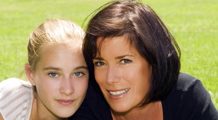 blond teenage girl and brunette mother smile on grassy area