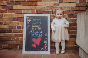 toddler standing next to sign with text born: date, adopted: date