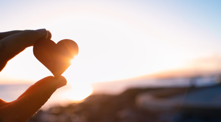 hand holding up small heart shaped stone against sunrise