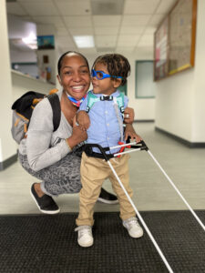 toddler using belt cane smiles at his mom who is kneeling next to him